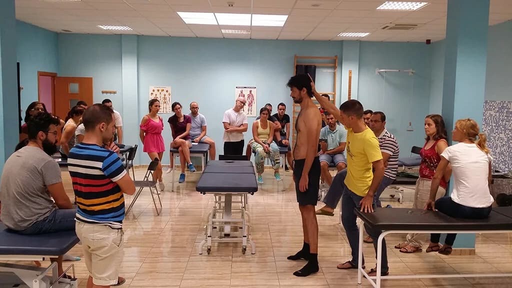 Uma turma de Manipulação Fascial à volta de um formando de pé, com uma pessoa a testar-lhe o movimento do ombro